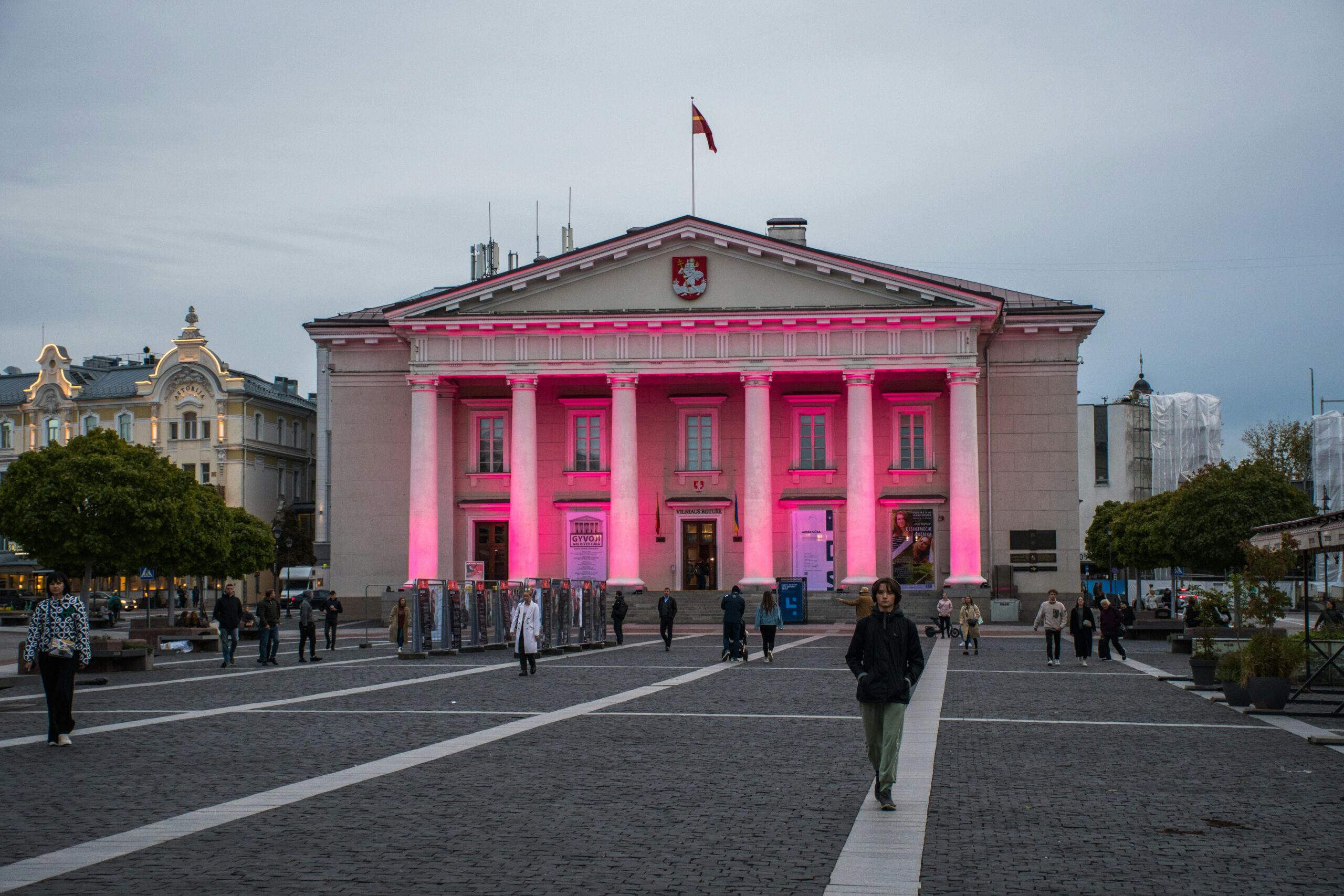 A peaceful evening scene at Vilnius Town Hall with people strolling and vibrant pink lighting.